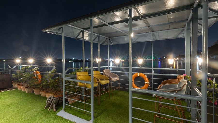 A rooftop seating area at night illuminated by overhead lights, with a river in the background and a lifebuoy hanging on the railing.