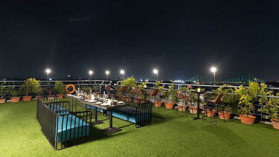 Nighttime view of a rooftop dining space with green flooring and table settings, with a bridge and river illuminated in the background.