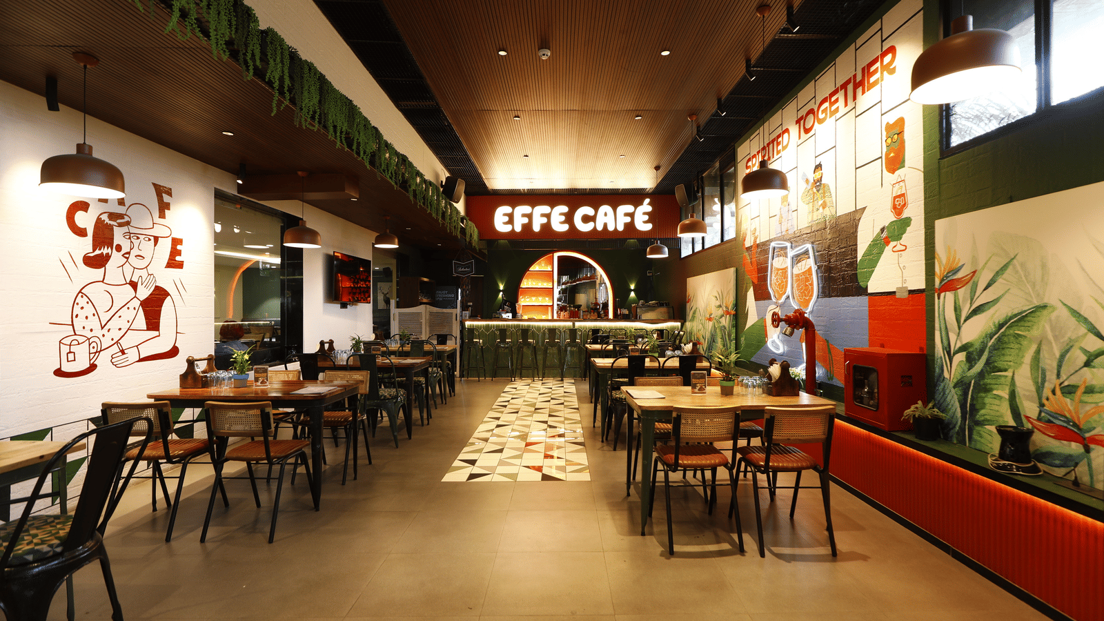 An Interior view of a modern cafe with wooden tables and chairs, green plants hanging from the ceiling, colorful wall murals, and a sign reading Effe Cafe at the back in Papaya Tree Hotels.