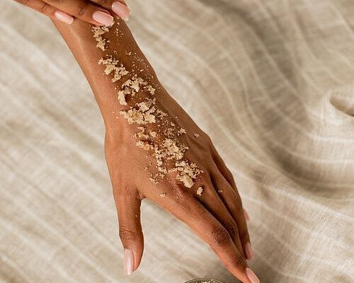 Woman applying a scrub or body polish to her forearm, with a jar of product placed nearby on beige fabric.