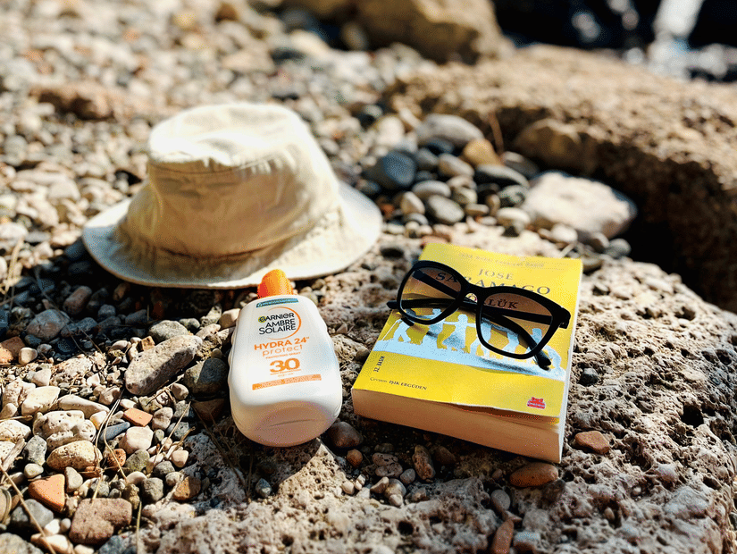 A straw hat, suncream bottle, and a book resting on a pebbled beach shoreline, with a bright blue sea and sky in the background.