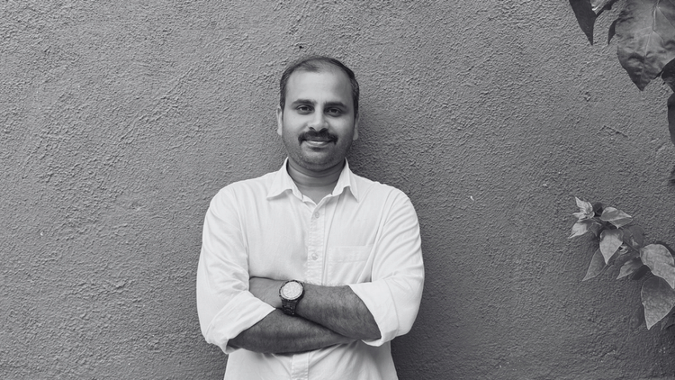 Dr. Rafeek Jabbar standing against a textured outdoor wall with arms crossed, wearing a light-colored shirt and wristwatch, next to leafy branches in the upper right corner.