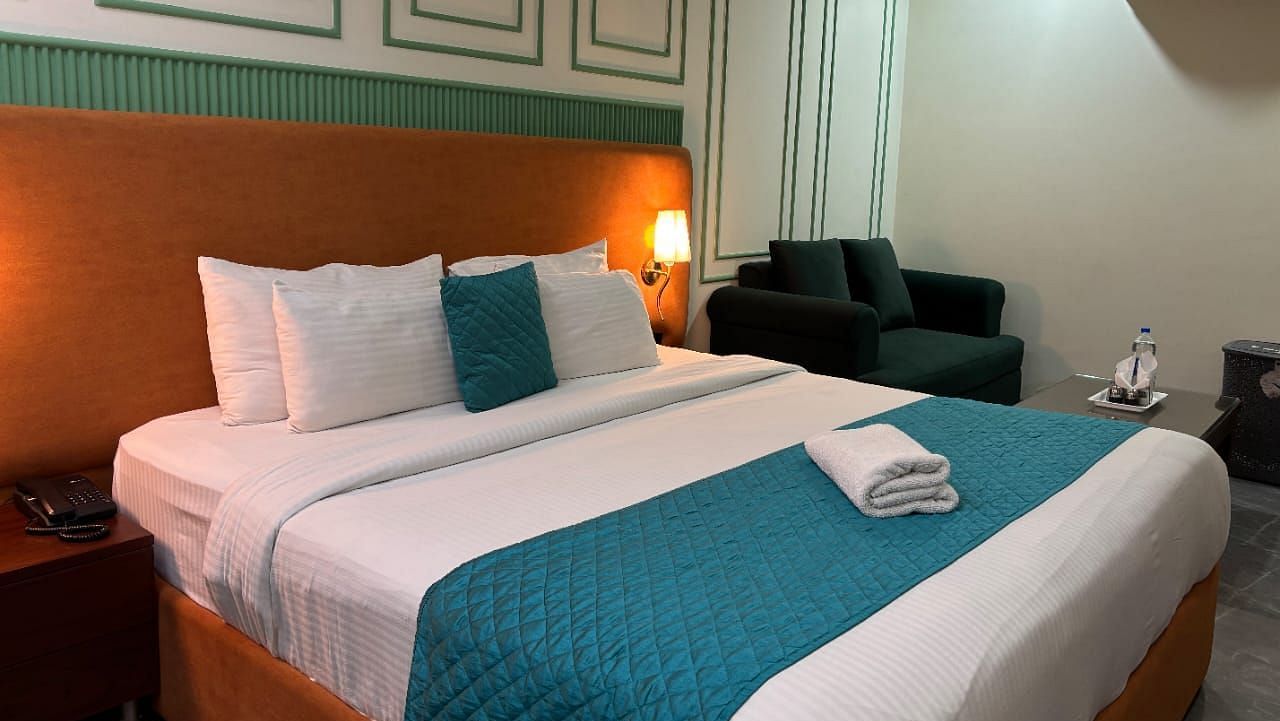 A well-lit room with a double bed dressed in white and blue, and a dark armchair - Asian Suites IFFCO Metro, Gurgaon