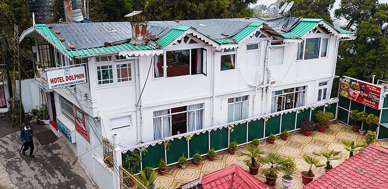 An aerial view of a multi-level white hotel building with green and red roofs nestled among trees on a hillside at Hotel Dolphin Darjeeling.