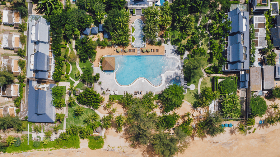 Aerial view of Natai Beach Resort featuring the spacious beachfront resort layout with a curved swimming pool, villas, and palm trees right next to the ocean.