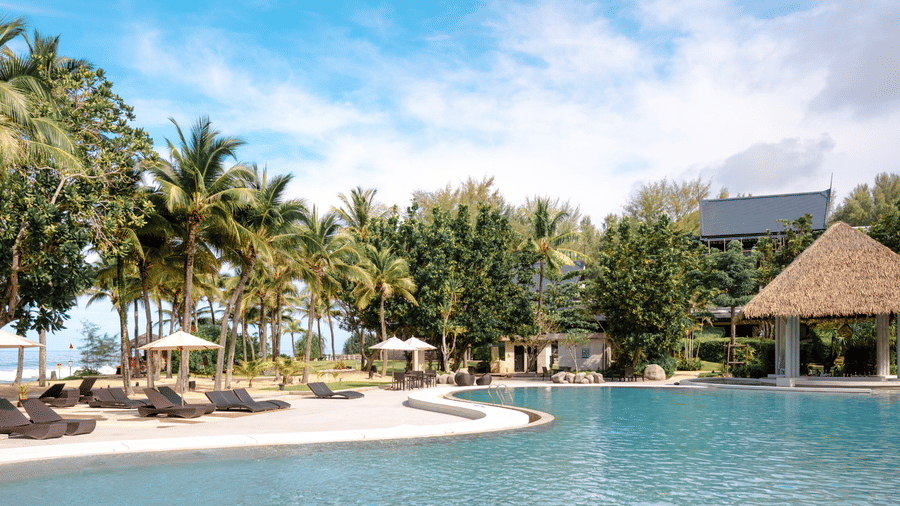 The large outdoor swimming pool at Natai Beach Resort surrounded by comfortable lounge chairs, umbrellas, tall palm trees, and a thatched roof pavilion.