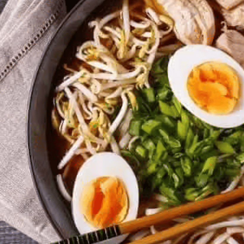 Bowl of ramen with soft-boiled eggs, noodles, sliced green onions, bean sprouts, and meat, with chopsticks placed across the bowl.