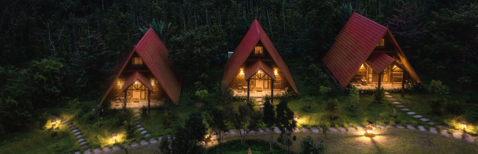 A nighttime aerial view of illuminated A-frame cottages nestled in the forest at The Copper Edge - Eco Resort, Coorg.