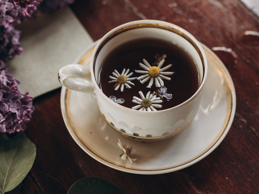 A cup of chamomile tea with chamomile flowers floating in it, on a saucer beside lilac blossoms and green leaves on a dark wooden table