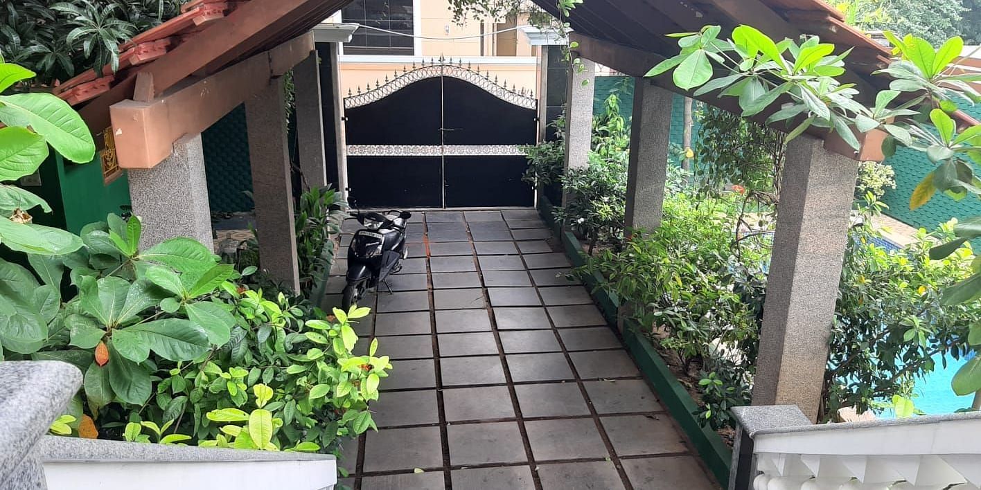 Covered outdoor walkway with a tiled path, surrounded by lush plants, leading to a gated entrance under a wooden roof structure.