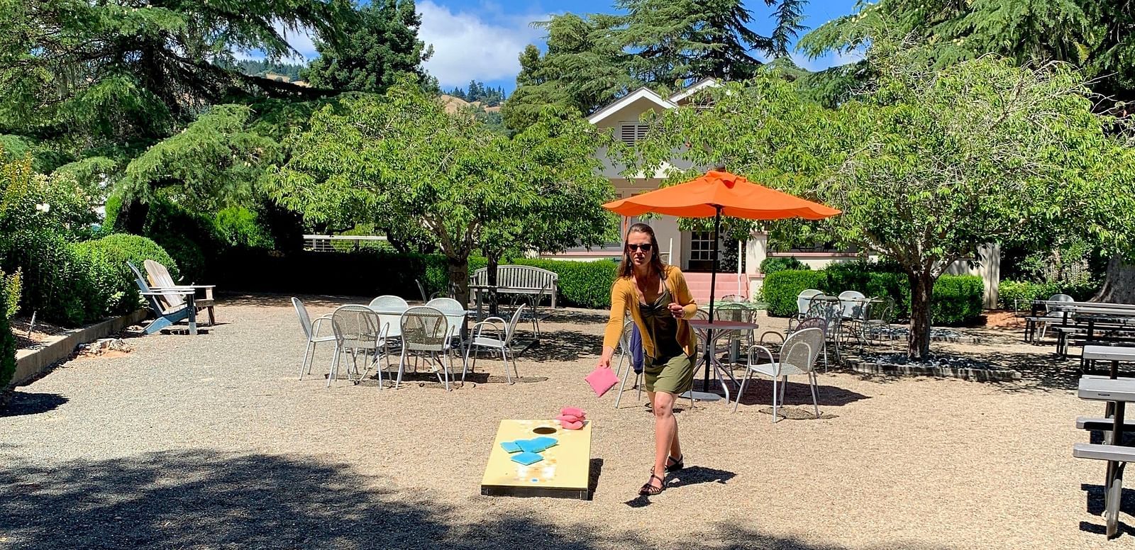 A person plays a beanbag toss game in a courtyard with trees, tables, and an umbrella.