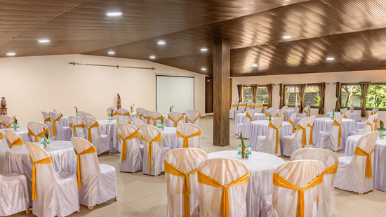 A large hall features round tables covered with white cloths and gold bands, sitting under a polished wooden ceiling with recessed lighting and plain beige walls.