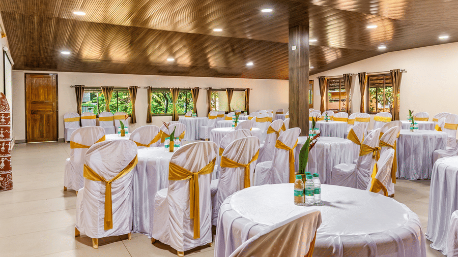 The event space contains many round tables dressed in white covers and gold bows, situated beneath a brown wooden ceiling fitted with small round bright lights.