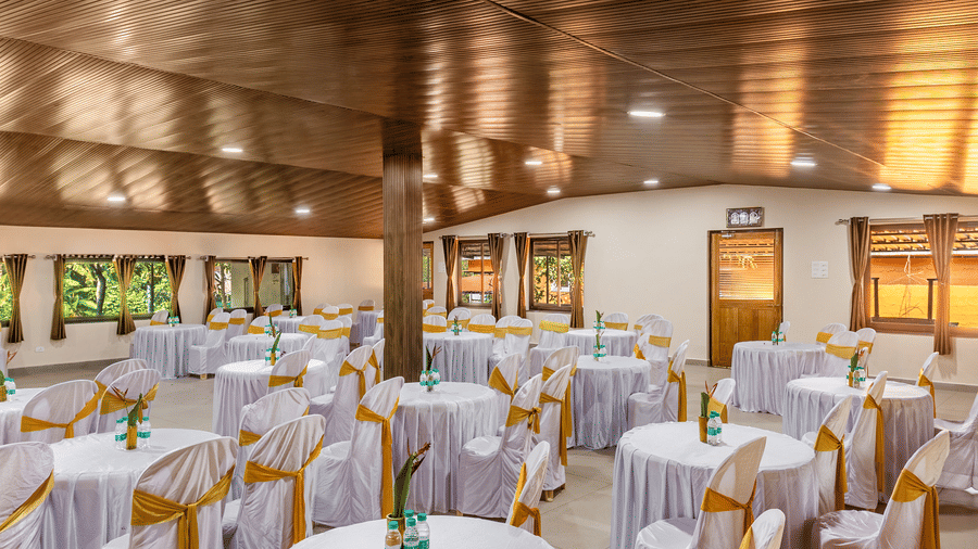 A view of the hall displays rows of tables dressed in white linens, set beneath a ceiling lined with wood panels and lights, with curtains on the back windows.