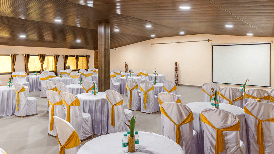 The spacious hall is arranged with round tables and white chairs for an event, featuring a large white projector screen standing at the far end of the big room.