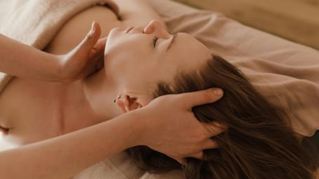  A client with her eyes closed lies on a massage table as a therapist gently massages her neck and head.