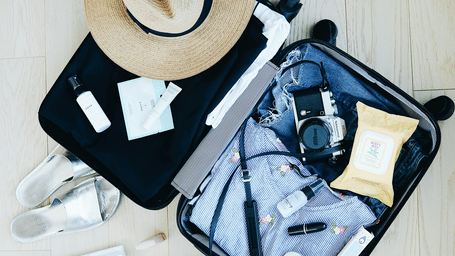 Open suitcase packed with clothes, a sun hat, camera, toiletries, sandals, and travel essentials arranged on a light wooden floor.