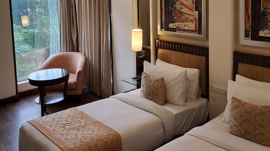 An image of a bedroom featuring twin bed, a sofa chair, a small centre table, bedside table with lamp, photo frames hanged on the wall, and a window o=overlooking the outdoors - Inde Hotel Signature Tower Gurgaon