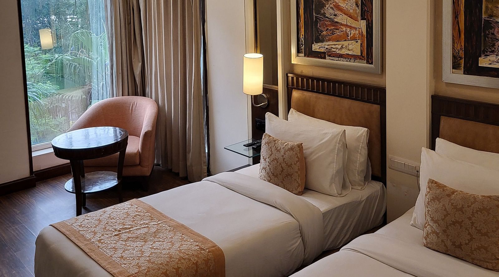 An image of a bedroom featuring twin bed, a sofa chair, a small centre table, bedside table with lamp, photo frames hanged on the wall, and a window o=overlooking the outdoors - Inde Hotel Signature Tower Gurgaon