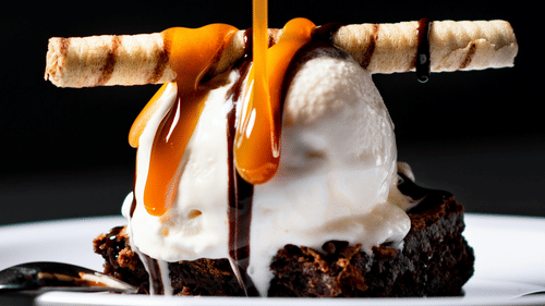 A dessert with ice cream on a brownie base topped with a chocolate stick and sauce being poured over.