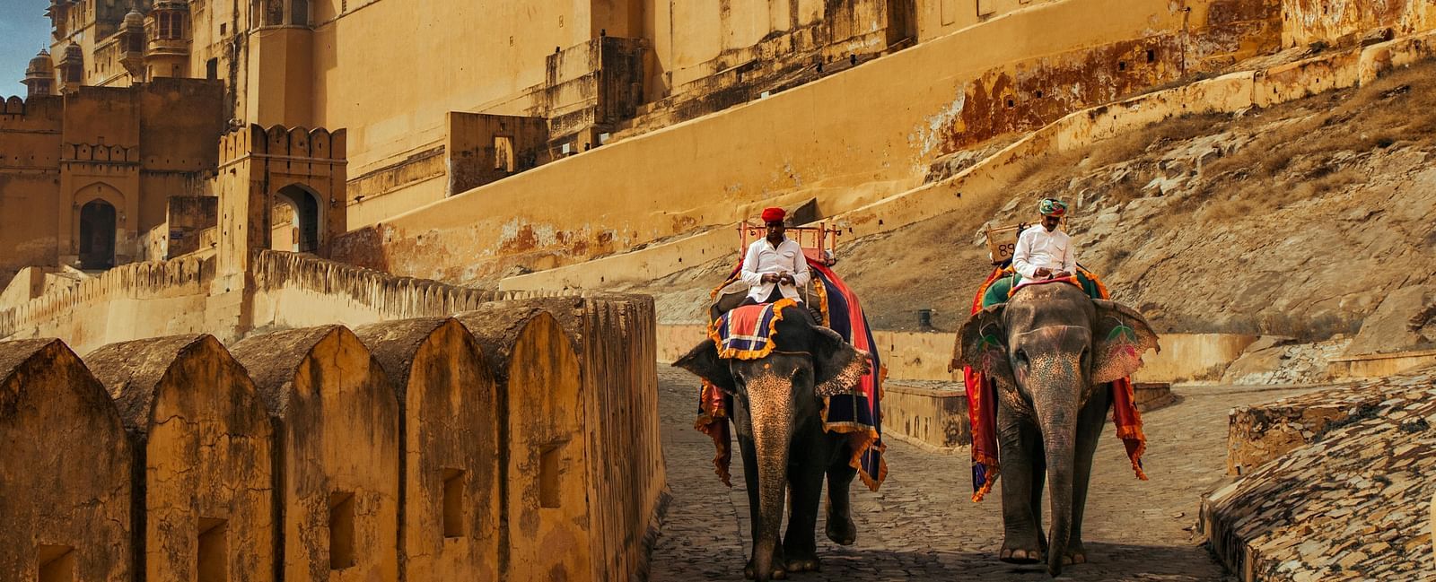 Two elephants carry tourists up a path towards the majestic Amber Fort in Jaipur, Rajasthan, under a partly cloudy sky.