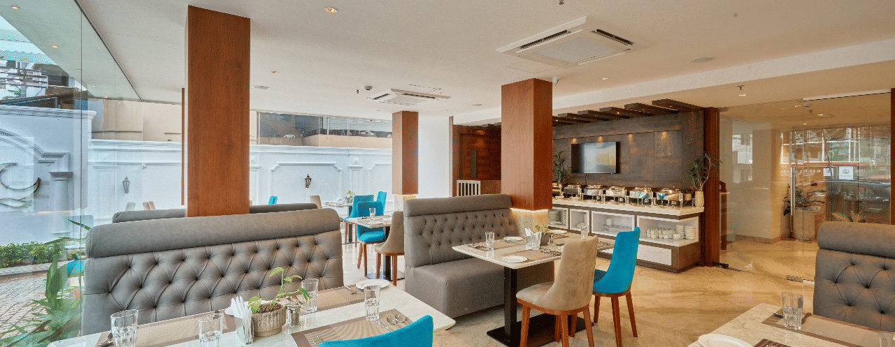 Wide view of the restaurant at QWhite Crescent Luxury Hotel by Dimora Kochi with teal chairs, cushioned booths, and warm ambient lighting.