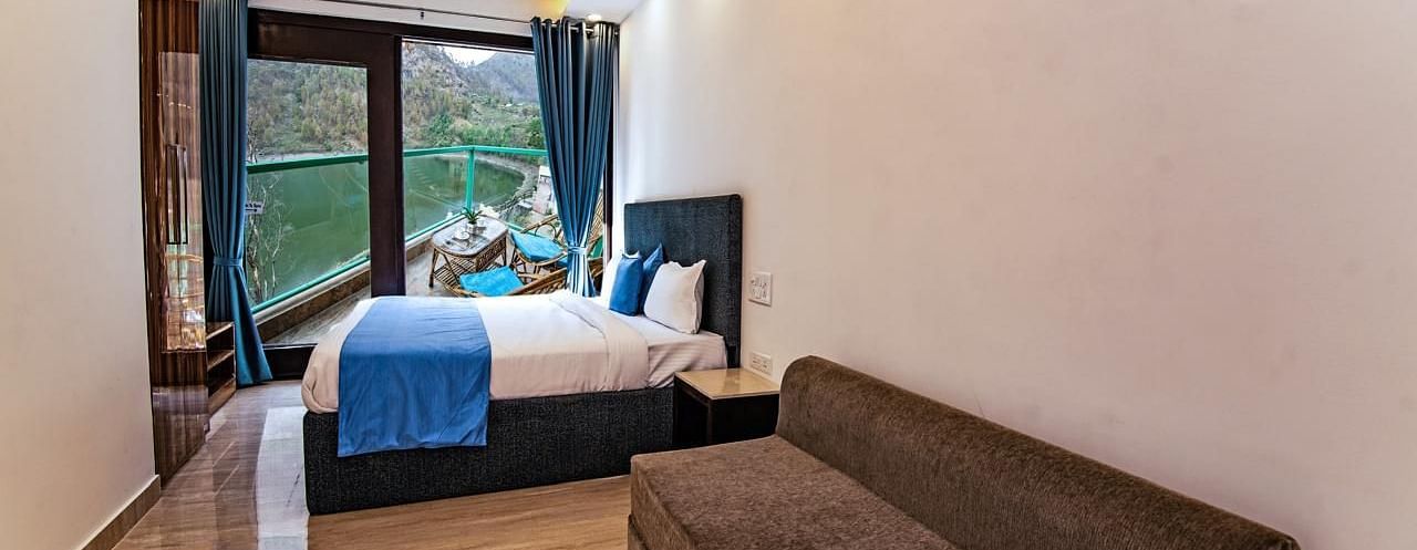 A bedroom with a large bed, sofa, wooden flooring, and full-length windows overlooking greenery.