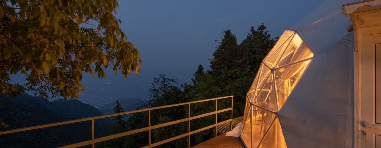 An image of wooden filled with warm lights emitting from a dome style cottage with the backdrop of evening sky filled with clouds -   Avalon Cottages, Kanatal