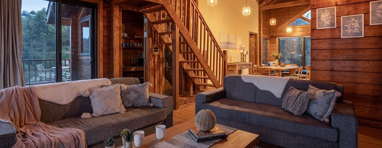 An image of a seating area with sofa set, centre table, frames hanged on wooden walls, and an indoor stair leading to next floor | Avalon Cottages, Kanatal