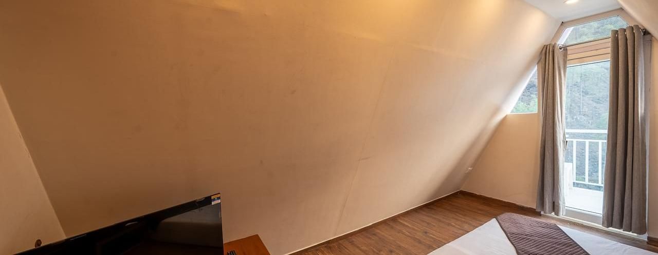 A modern hotel room with sloped light-coloured walls, a bed, a television on a stand, and a large window to the right.