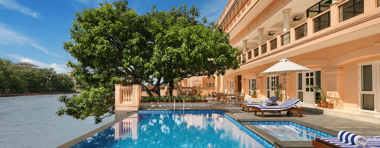 Outdoor swimming pool beside a river with lounge chairs and umbrellas, next to a peach-coloured hotel building with balconies and a tree providing shade - Auradoon, Dehradun