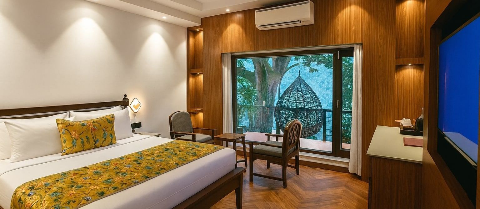 Premium Ganges View Room at Summit by the Ganges Beach Resort and Spa, Rishikesh featuring a double size bed, a sitting area with chairs and centre table, a cabinet table, a flat TV screen, an AC, and a balcony attached which has a chair placed.