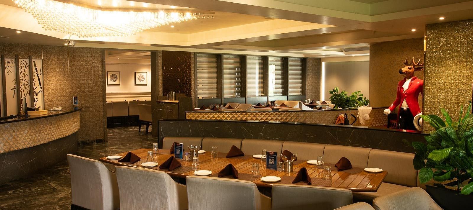 Dining area features warm, welcoming lighting, a large table surrounded by cushioned seating, and a tall deer-faced statue complementing the cheerful setting  | Crescent Spa & Resorts