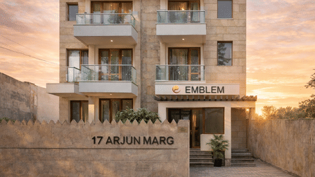 Exterior facade of Emblem Hotel at 17 Arjun Marg Gurgaon featuring a modern sandstone building with balconies, branded signage, and a warm sunset sky in the background.