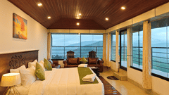 The well-lit suite mountain view at Ibex Resorts, Valparai (Tale Rock), featuring  a large bed, bedspreads, pillows, a sofa, and large windows with greenery in the backdrop.