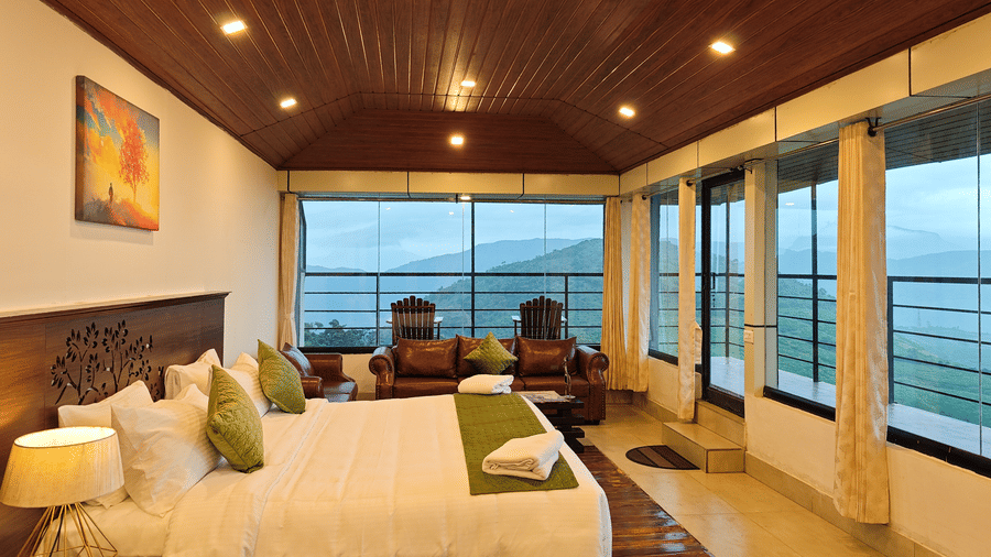 The well-lit suite mountain view at Ibex Resorts, Valparai (Tale Rock), featuring  a large bed, bedspreads, pillows, a sofa, and large windows with greenery in the backdrop.