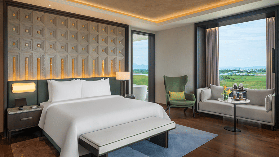 Executive Suite main bedroom with a double bed, padded bench, green armchair and a sofa by a window looking over countryside at The Residency Signature Madurai.