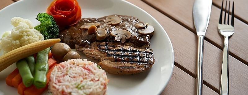 A plate featuring grilled meat, rice salad, mashed potatoes, steamed vegetables, and a tomato rose garnish, with cutlery placed beside it on a wooden table at The Hamuse Hotel, Kodaikanal.