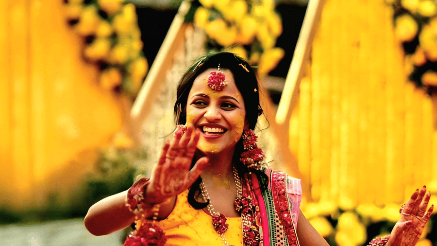 A bride in a red and yellow traditional outfit sitting outdoors at Vedic Village Spa Resort surrounded by marigold floral decorations and a thatched structure in the background.