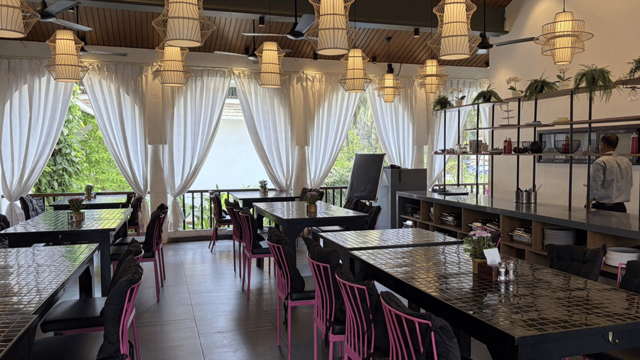 Rows of dining tables, pink chairs, and hanging lanterns fill a large restaurant with draped windows at Bougainvillea, Alibaug.