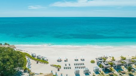 An aerial view of a resort beach with white sand, blue ocean, numerous lounge setups, and thatched structures.