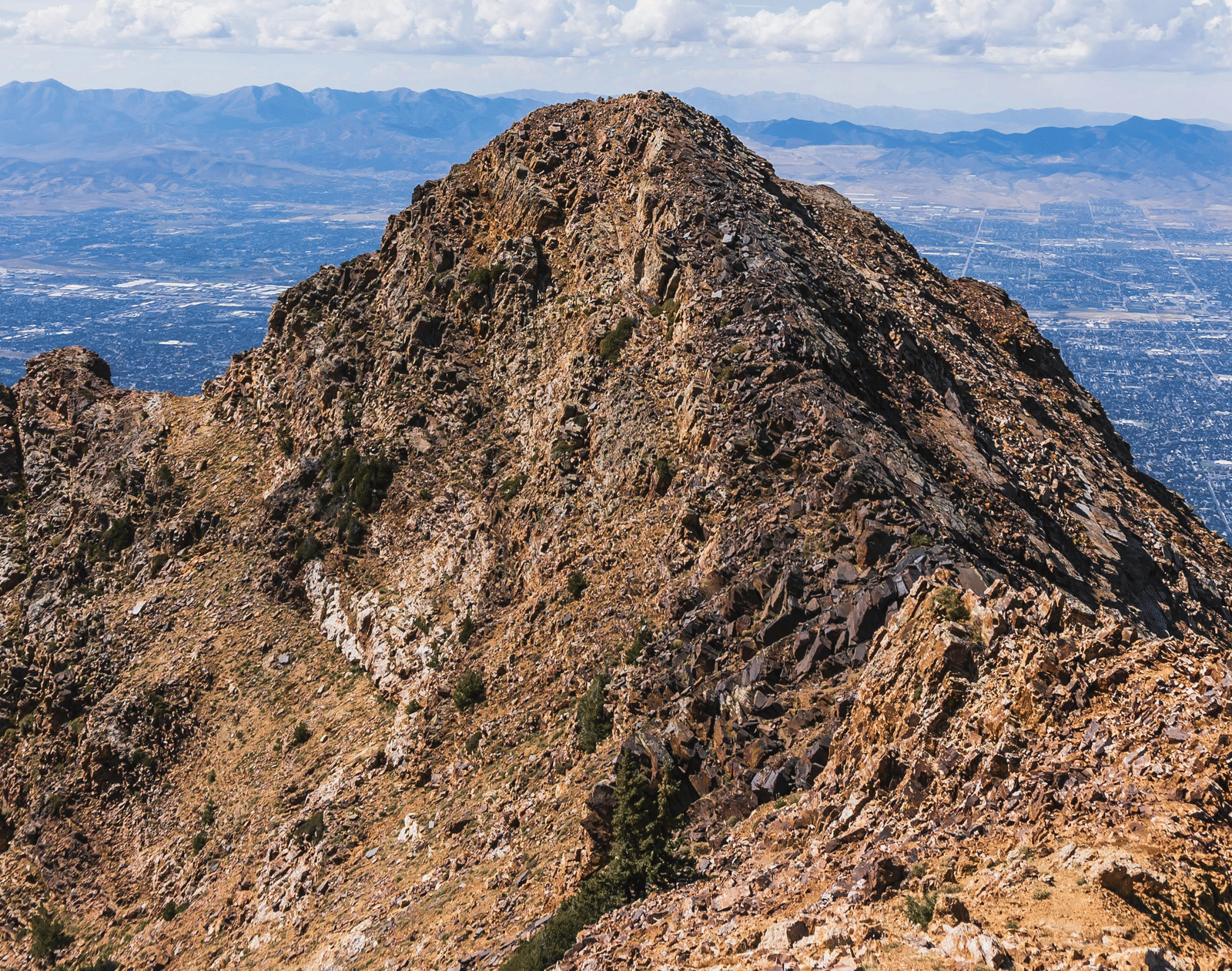 A rugged, barren mountain peak under a blue sky with white clouds. The peak is composed of rocky, brown terrain and appears as a sharp, triangular summit in the foreground.