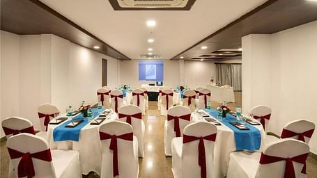 A banquet hall setup with round tables covered in white cloths and chairs adorned with bows, under ceiling lights at Lords Eco Inn Jayanagar Bangalore.