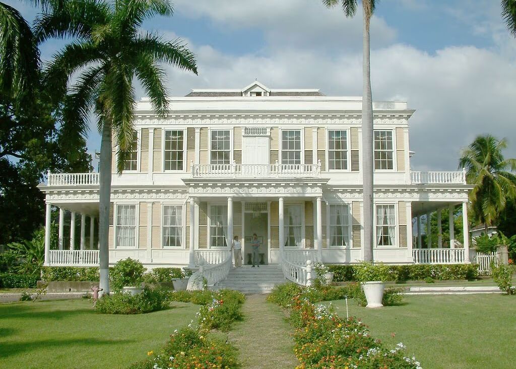 A grand white colonial-style building with columns and multiple windows stands amidst a manicured green lawn with palm trees.