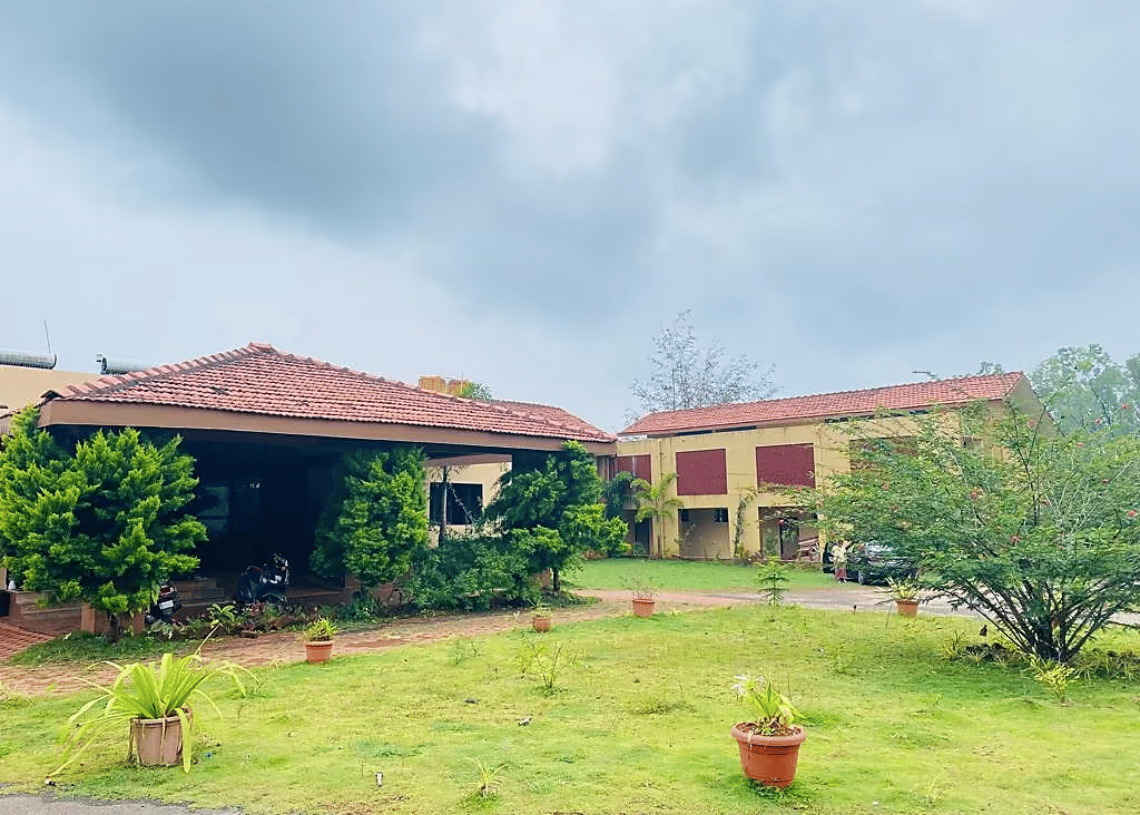 Building with tiled roof and green plants in front, surrounded by grass and potted plants on a cloudy day at  Paradise Wild Hills Resort, Kollur.