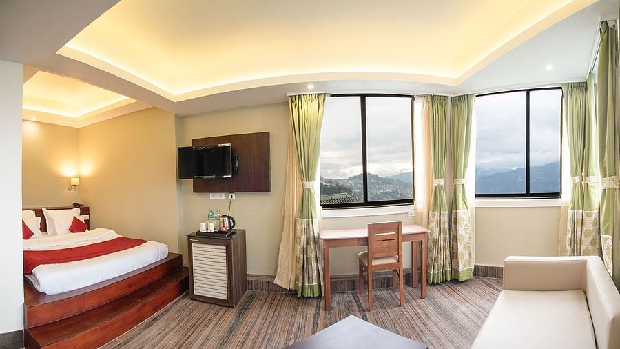 A panoramic view of a room featuring a bed, a seating area, a wall-mounted television, and a window with a view - Summit North Star, Gangtok