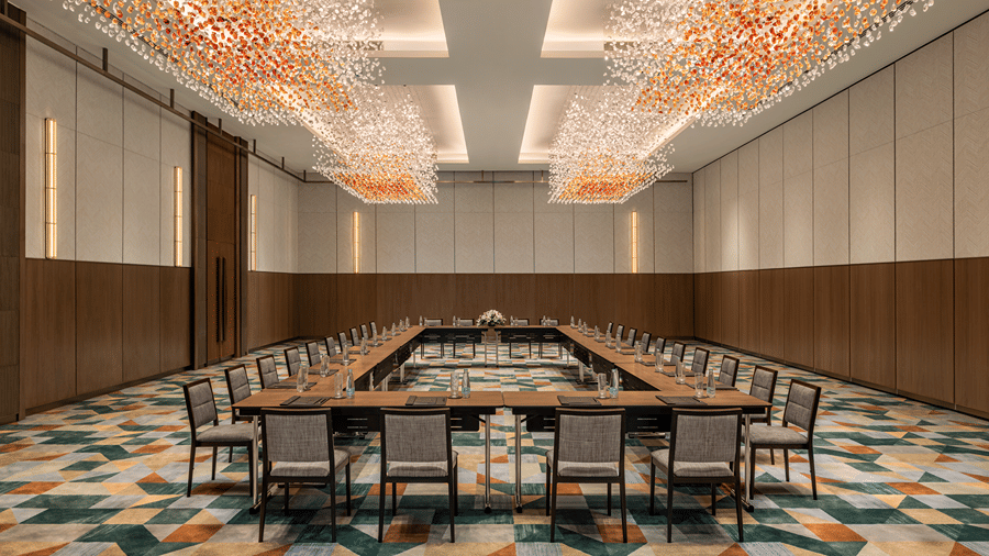The large meeting room of Ballroom 3 configured in board room arrangement with warm crystal chandeliers and colourful patterned carpet at The Residency Towers Chennai.