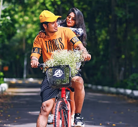A man riding a bicycle and a woman sitting behind him, looking at each other with flowers in the cycle basket, surrounded by greenary