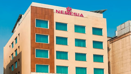 Daytime view of the Nemesia hotel building facade with teal-tinted windows, wooden accents, and palm trees in front.