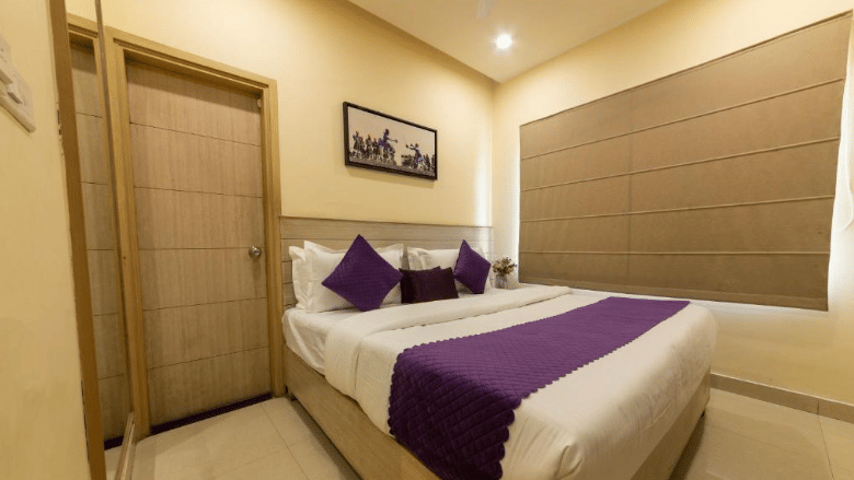 A hotel room, featuring a bed with white linens and a purple runner and pillows, a wooden door, and a horizontal window blind | Perfectstayz Value Sagar